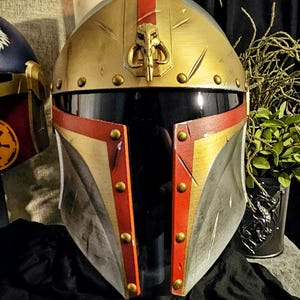 May include: A gold, red, and silver Mandalorian helmet with a T-shaped visor. The helmet features a gold crest with a skull emblem and is adorned with gold rivets. The design is reminiscent of a popular science fiction franchise.