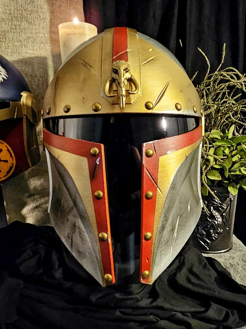 May include: A gold, red, and silver Mandalorian helmet with a T-shaped visor. The helmet features a gold crest with a skull emblem and is adorned with gold rivets. The design is reminiscent of a popular science fiction franchise.