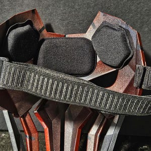 K&ouml;nnte beinhalten: Nahaufnahme einer rot-silbernen Maske mit schwarzer Polsterung und einem schwarzen, verstellbaren Riemen. Die Maske hat ein geometrisches Design und ist wahrscheinlich f&uuml;r Cosplay oder Kost&uuml;me gedacht.