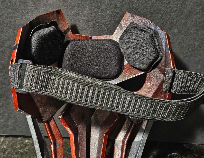 K&ouml;nnte beinhalten: Nahaufnahme einer rot-silbernen Maske mit schwarzer Polsterung und einem schwarzen, verstellbaren Riemen. Die Maske hat ein geometrisches Design und ist wahrscheinlich f&uuml;r Cosplay oder Kost&uuml;me gedacht.