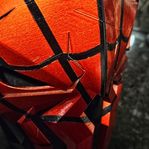 K&ouml;nnte beinhalten: Nahaufnahme eines rot-schwarzen Objekts mit strukturierter Oberfl&auml;che. Das Objekt hat ein geometrisches Design mit schwarzen Linien, die ein spinnennetzartiges Muster bilden. Die rote Farbe hat einen metallischen Glanz, und das Objekt scheint 3D-gedruckt zu sein.