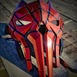 K&ouml;nnte beinhalten: Eine rot-schwarze Maske mit geometrischen Designs. Die Maske hat eine spitze Form mit kantigen Merkmalen und Augen&ouml;ffnungen. Schwarze Linien und Akzente sind im gesamten Design sichtbar. Die Maske besteht aus einem harten Material.