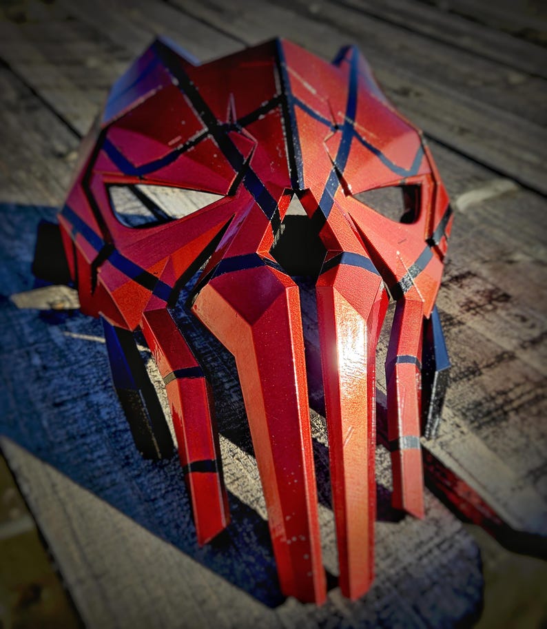 K&ouml;nnte beinhalten: Eine rot-schwarze Maske mit geometrischen Designs. Die Maske hat eine spitze Form mit kantigen Merkmalen und Augen&ouml;ffnungen. Schwarze Linien und Akzente sind im gesamten Design sichtbar. Die Maske besteht aus einem harten Material.