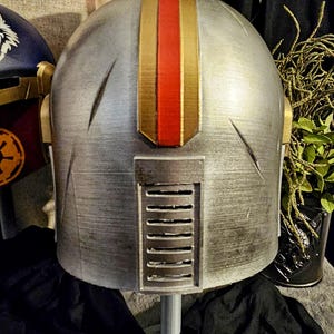 May include: A silver helmet with gold and red accents. The helmet has a vertical gold stripe with a red center stripe. The back of the helmet features a rectangular vent. The helmet is part of a costume or cosplay.