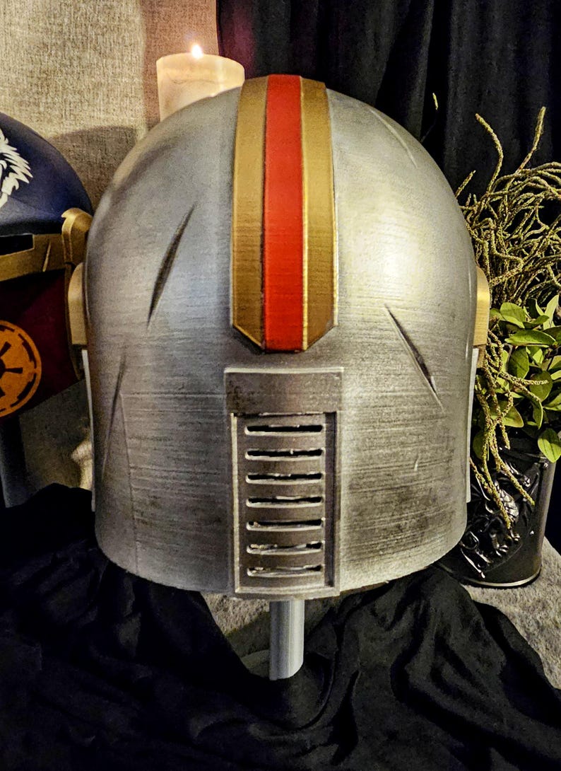 May include: A silver helmet with gold and red accents. The helmet has a vertical gold stripe with a red center stripe. The back of the helmet features a rectangular vent. The helmet is part of a costume or cosplay.