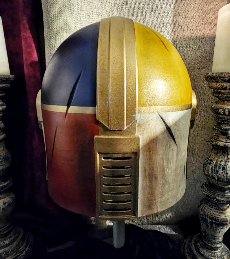 May include: A custom-painted helmet with a segmented design. The top is split between blue and yellow, with a gold central ridge. The lower sections are red and off-white, with gold accents and a vent detail. A unique cosplay item.