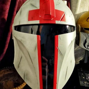 May include: A white and gray helmet with a red cross design on top and red stripes down the front. The helmet has a black visor and appears to be a prop or costume piece. The overall design suggests a futuristic or sci-fi theme.