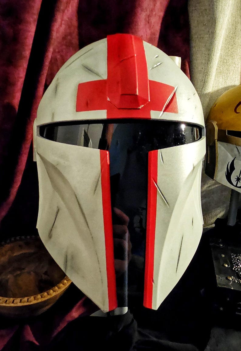 May include: A white and gray helmet with a red cross design on top and red stripes down the front. The helmet has a black visor and appears to be a prop or costume piece. The overall design suggests a futuristic or sci-fi theme.