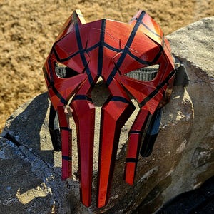 K&ouml;nnte beinhalten: Eine rot-schwarze geometrische Maske mit auff&auml;lligem Design. Die Maske weist eine Reihe von eckigen Formen und Linien auf, die eine k&uuml;hne und futuristische &Auml;sthetik erzeugen. Die Maske besteht aus einem harten Material und hat ein gl&auml;nzendes Finish.