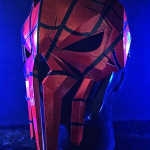 K&ouml;nnte beinhalten: Eine rot-schwarze Maske mit geometrischem Design. Die Maske hat ein facettiertes Aussehen mit scharfen Winkeln und Linien. Die Maske wird auf einer schwarzen Kopfattrappe vor blauem Hintergrund pr&auml;sentiert.