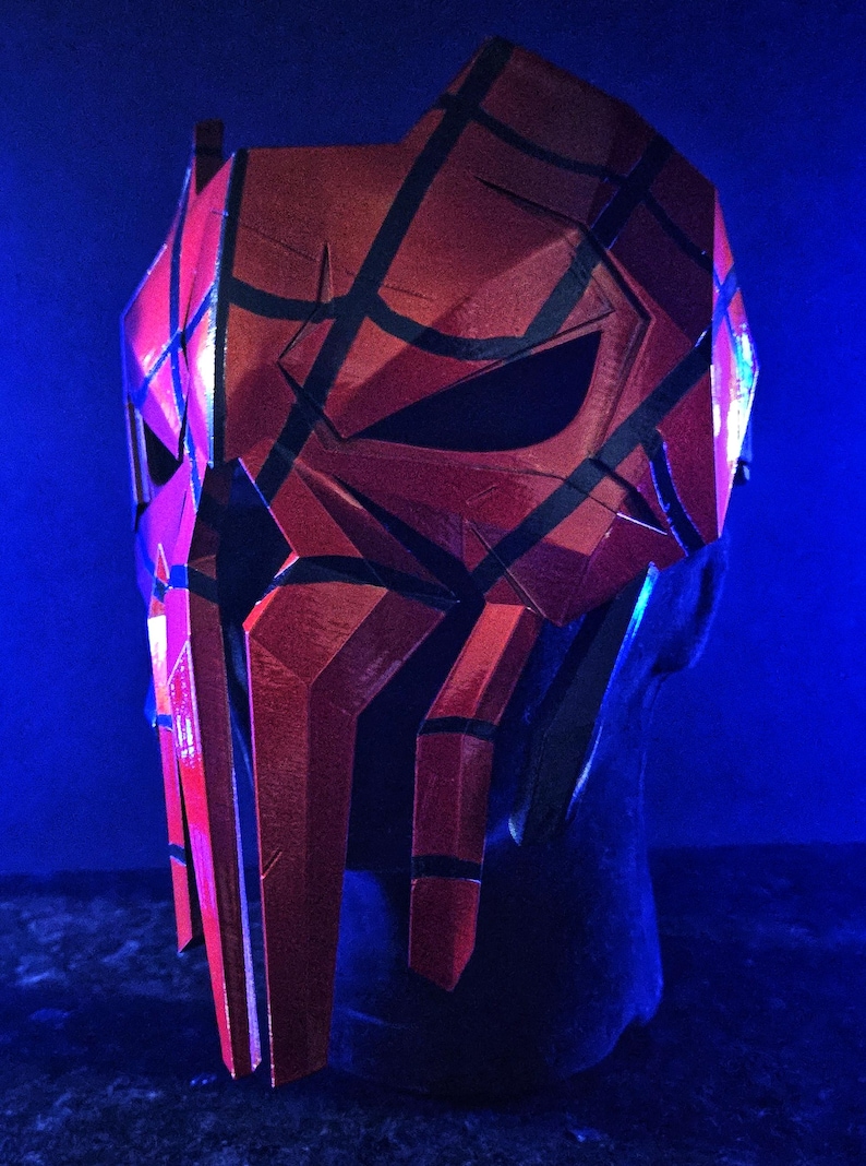 K&ouml;nnte beinhalten: Eine rot-schwarze Maske mit geometrischem Design. Die Maske hat ein facettiertes Aussehen mit scharfen Winkeln und Linien. Die Maske wird auf einer schwarzen Kopfattrappe vor blauem Hintergrund pr&auml;sentiert.