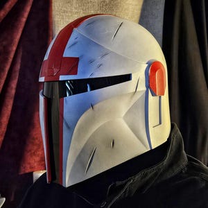 May include: A white and gray helmet with red accents, featuring a visor and a distinctive cross design on top. The helmet has visible wear and tear marks, suggesting a battle-worn aesthetic. The design is reminiscent of a sci-fi or fantasy theme.