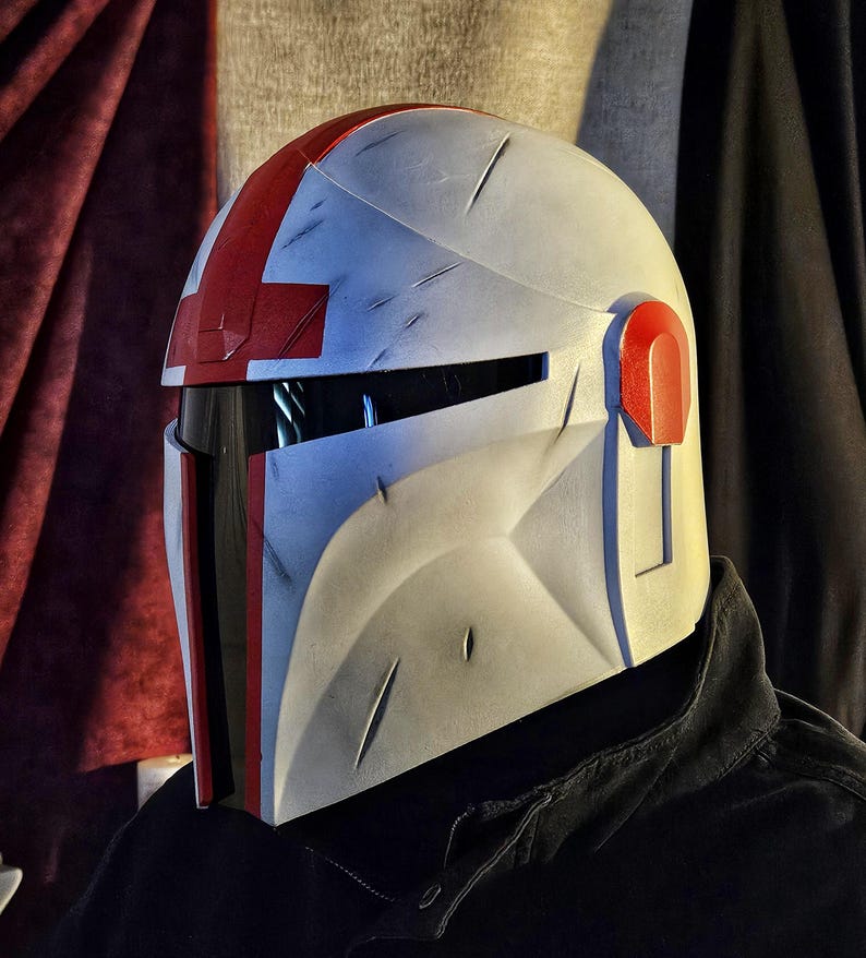 May include: A white and gray helmet with red accents, featuring a visor and a distinctive cross design on top. The helmet has visible wear and tear marks, suggesting a battle-worn aesthetic. The design is reminiscent of a sci-fi or fantasy theme.