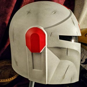 May include: A weathered, off-white helmet with red accents. The helmet features a visor and is designed with angular shapes and visible wear marks. The design includes a red ear piece and a dark visor. The helmet is likely for cosplay or costume use.