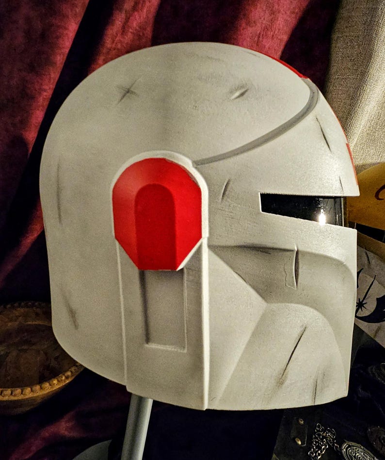 May include: A weathered, off-white helmet with red accents. The helmet features a visor and is designed with angular shapes and visible wear marks. The design includes a red ear piece and a dark visor. The helmet is likely for cosplay or costume use.