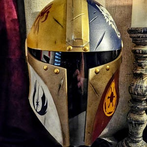 May include: A custom-painted Mandalorian helmet with a gold, blue, and red color scheme. The helmet features various symbols and designs, including a spider, a bird, and a starburst. The visor is black, and the helmet is adorned with gold rivets.