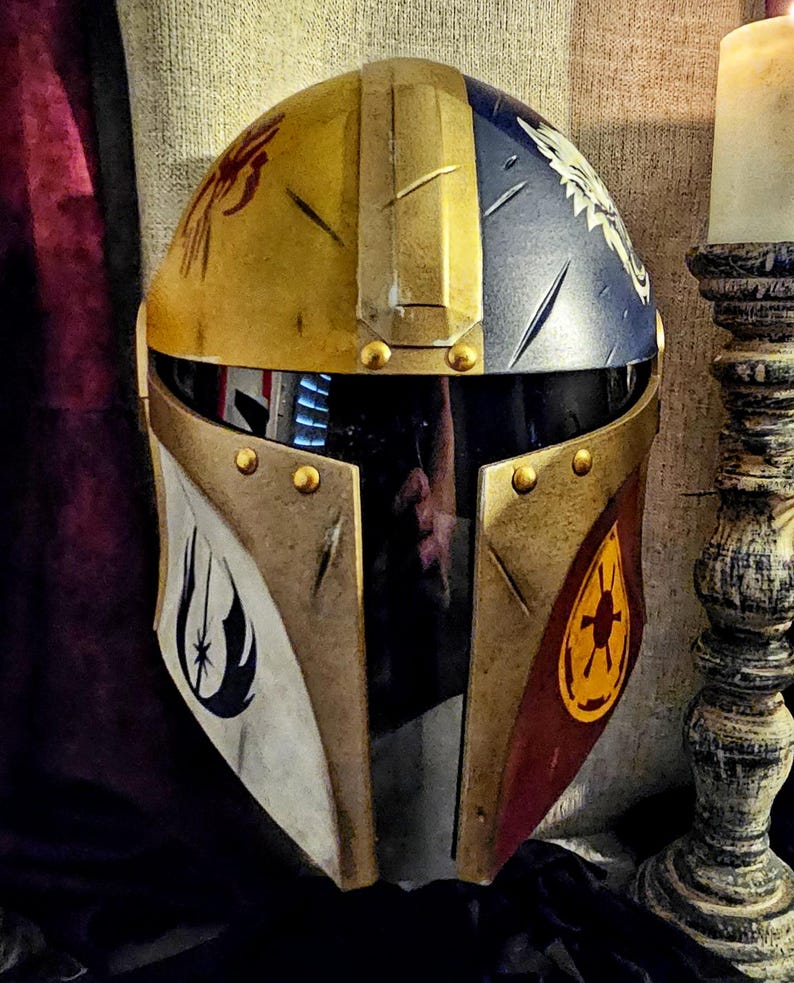 May include: A custom-painted Mandalorian helmet with a gold, blue, and red color scheme. The helmet features various symbols and designs, including a spider, a bird, and a starburst. The visor is black, and the helmet is adorned with gold rivets.