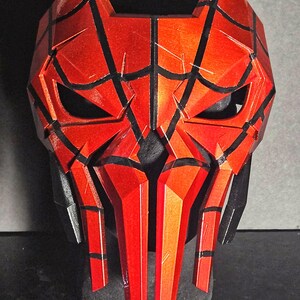 K&ouml;nnte beinhalten: Eine rot-schwarze Maske mit geometrischem Design. Die Maske hat eckige Merkmale, einschlie&szlig;lich spitzer Kanten und eines segmentierten Aussehens. Die Augen&ouml;ffnungen sind dunkel, und die Maske ist vor einem dunklen Hintergrund platziert.