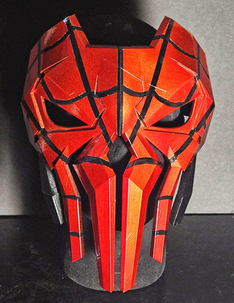 K&ouml;nnte beinhalten: Eine rot-schwarze Maske mit geometrischem Design. Die Maske hat eckige Merkmale, einschlie&szlig;lich spitzer Kanten und eines segmentierten Aussehens. Die Augen&ouml;ffnungen sind dunkel, und die Maske ist vor einem dunklen Hintergrund platziert.