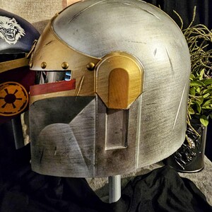 May include: A metallic silver and gold helmet with a visor and a rectangular faceplate. The helmet has a weathered appearance with visible scratches and rivets. The design includes gold accents and a red stripe near the visor.
