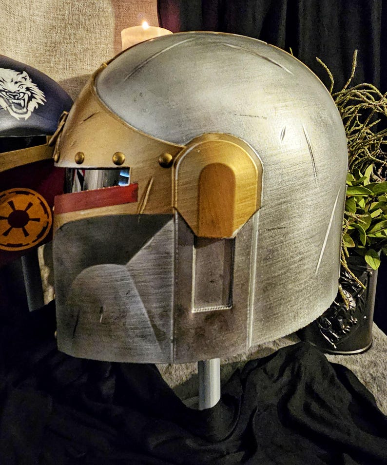 May include: A metallic silver and gold helmet with a visor and a rectangular faceplate. The helmet has a weathered appearance with visible scratches and rivets. The design includes gold accents and a red stripe near the visor.