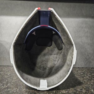 May include: An inside view of a helmet with a gray interior and white trim. The helmet has a black padded interior and a blue and red band. The helmet is designed to protect the head.