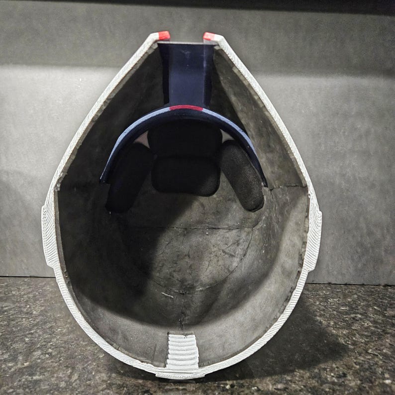 May include: An inside view of a helmet with a gray interior and white trim. The helmet has a black padded interior and a blue and red band. The helmet is designed to protect the head.