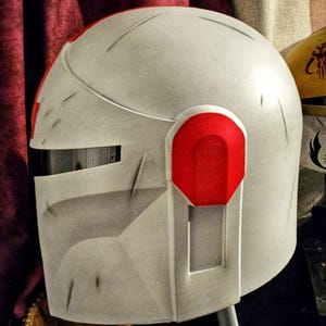May include: A white and red helmet with a visor and ear pieces. The helmet has a weathered appearance with visible scratches and a red accent on the ear piece. The design is reminiscent of a futuristic or science fiction aesthetic.