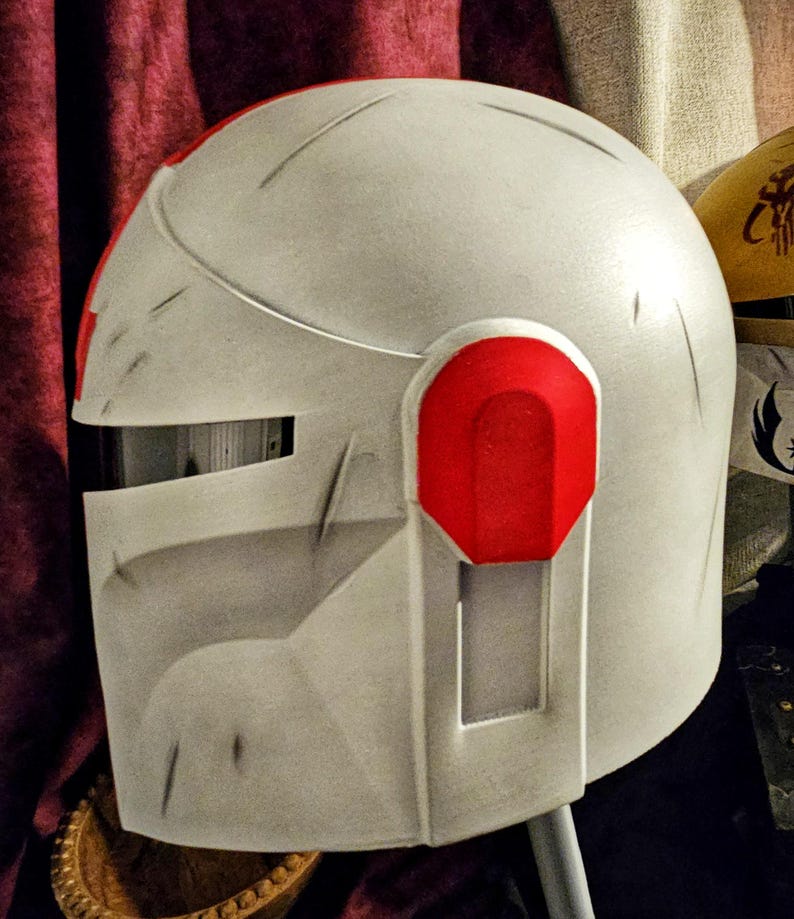 May include: A white and red helmet with a visor and ear pieces. The helmet has a weathered appearance with visible scratches and a red accent on the ear piece. The design is reminiscent of a futuristic or science fiction aesthetic.