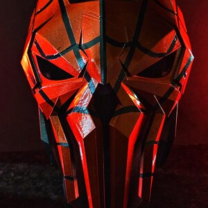 K&ouml;nnte beinhalten: Eine geometrische, rot-schwarze Maske mit eckigen Formen und Augen&ouml;ffnungen. Die Maske hat eine strukturierte Oberfl&auml;che und einen dunklen Hintergrund, wodurch ein dramatischer Effekt entsteht. Das Design ist symmetrisch und scheint handgefertigt zu sein.