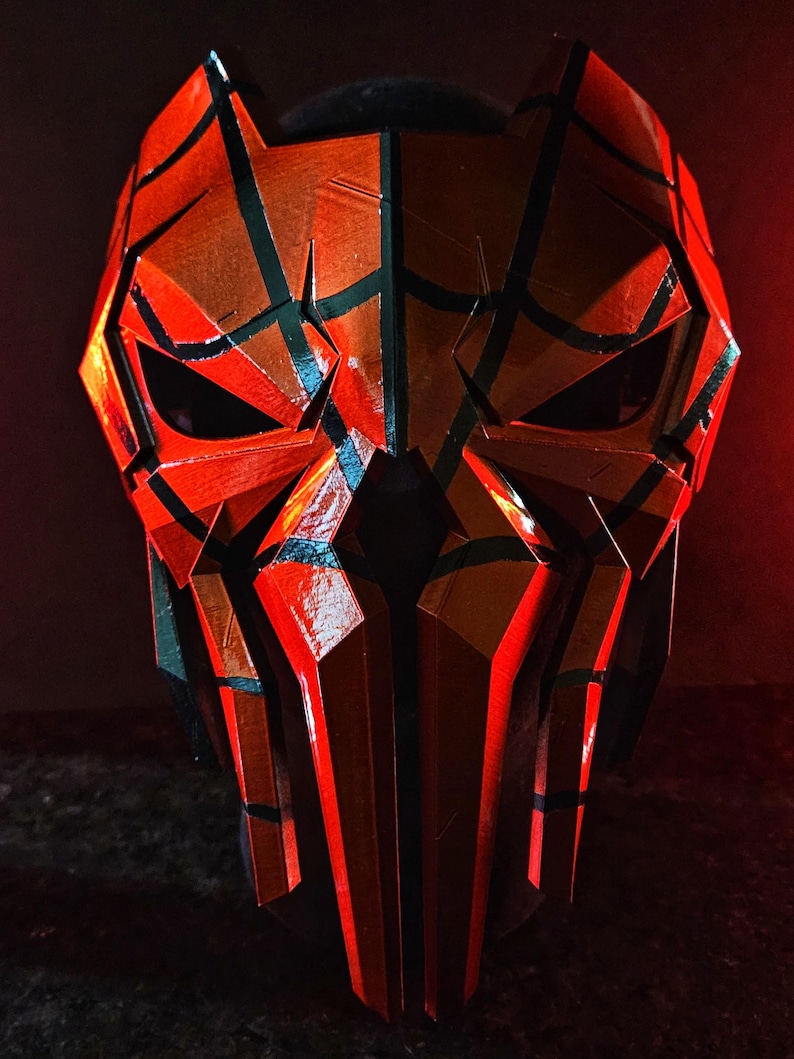 K&ouml;nnte beinhalten: Eine geometrische, rot-schwarze Maske mit eckigen Formen und Augen&ouml;ffnungen. Die Maske hat eine strukturierte Oberfl&auml;che und einen dunklen Hintergrund, wodurch ein dramatischer Effekt entsteht. Das Design ist symmetrisch und scheint handgefertigt zu sein.