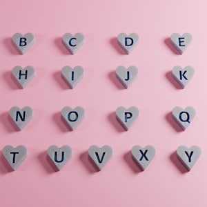 Puede incluir: Un conjunto de 26 letras del alfabeto en forma de corazones. Las letras están impresas en azul sobre un fondo gris claro. Los corazones están dispuestos en cuatro filas de seis corazones cada una. Los corazones están sobre un fondo rosa.
