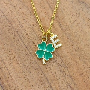 May include: Gold necklace with a green four-leaf clover charm and a letter 'E' charm. The charms are on a gold chain.