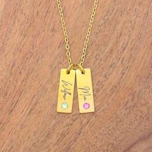 May include: A gold-toned necklace featuring two rectangular vertical pendants suspended from a delicate chain. Each pendant is engraved with a name in script, Willow and Mia, and is accented with a small, round, colored gemstone at the base.
