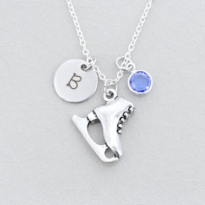 Op de afbeelding: Een zilveren ketting met een schaatsbedel, een ronde bedel met de letter "B" en een kleine blauwe edelsteen. De ketting hangt aan een zilveren ketting tegen een lichtgrijze achtergrond.