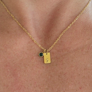 Op de afbeelding: Een goudkleurige ketting met een kleine rechthoekige hanger gegraveerd met de letter G en een aparte ronde bedel met een donkergroene steen. De fijne metalen ketting rust op de huid en benadrukt de twee kleine bedels.