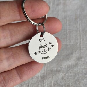 May include: A silver-colored keychain with a round pendant. The pendant features a cat face illustration, three small hearts, and the words "Cat Mom" in a simple font. The keychain is attached to a silver ring.