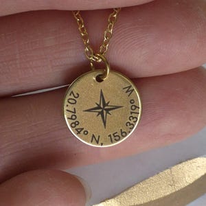 Puede incluir: Un collar con colgante dorado con un diseño de brújula. El colgante redondo tiene una rosa de los vientos en el centro y coordenadas alrededor del borde. El collar lo sostiene una persona.