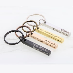 May include: A collection of personalized keychains in silver, gold, and black. Each rectangular bar is engraved with a message, including "Drive Safe," "Text Mom," and other phrases. The keychains are attached to metal rings.