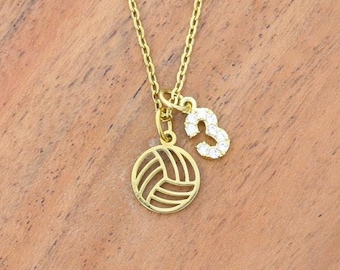 Può includere: Una collana color oro con una catenina sottile e due ciondoli: un pendente circolare a forma di pallavolo con linee traforate e un piccolo numero tre tempestato di pietre chiare e scintillanti che catturano la luce.