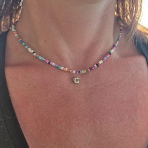 May include: A colorful beaded necklace with a small gold-colored pendant featuring the letter "G". The necklace is composed of small, multi-colored beads in shades of blue, pink, white, and orange. The necklace is worn around the neck.
