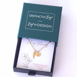 Könnte beinhalten: Goldfarbene Halskette mit Flügelanhänger und rundem Anhänger mit dem Buchstaben "K" in einer tealfarbenen Geschenkbox. Auf der Box steht "DISTINCTLY Ivy" und "Ivy BY DESIGN". Die Karte lautet "In Loving Memory".