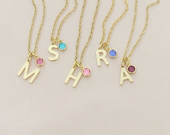 May include: Gold-colored initial necklaces with a delicate chain. Each necklace features a single letter pendant and a small, colorful gemstone charm. The letters are 'M', 'S', 'H', 'R', and 'A'. The gemstones are pink, blue, and purple.