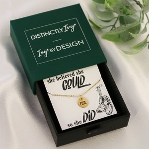 May include: A gold necklace with a round pendant that says "run." The necklace is in a green box with the text "Distinctly Ivy Ivy by Design." The box is on a white surface with a green plant in the background. The card inside the box says "She believed she could so she did."
