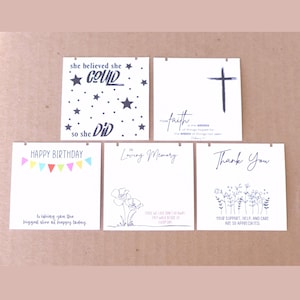 May include: A collection of white square cards with various messages and designs. One card reads "She believed she could so she did" with stars. Other cards say "Happy Birthday," "In Loving Memory," and "Thank You."
