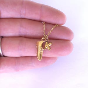 May include: A gold-colored necklace featuring a charm shaped like a cleat and a letter "K". The pendant hangs from a delicate gold chain. The cleat charm has detailed ridges and studs.