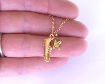 May include: A gold-coloured necklace with a charm shaped like a football boot and the letter "K". The pendant hangs from a fine gold chain. The boot charm has detailed ridges and studs.