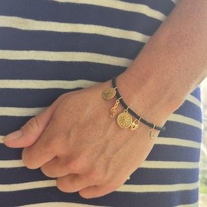May include: A black cord bracelet with gold charms. The charms include a compass, a musical note, a padlock, a small charm, and a charm with crossed golf clubs. The bracelet is on a wrist with a blue and white striped background.