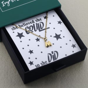 Op de afbeelding: Een goudkleurige metalen ketting met een letter M hanger, gepresenteerd in een donkergroene doos op een witte kaart met zwarte sterren en de tekst "she believed she COULD so she DiD" gedrukt in een vet, gestileerd zwart lettertype.