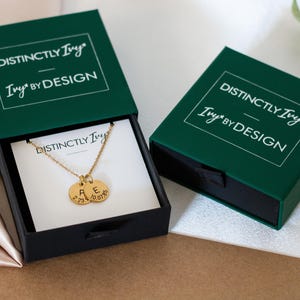 May include: A gold-toned necklace with two circular pendants featuring the letters A and E and dates 03.23.19 and 10.07.85, presented in a dark green sliding gift box labeled Distinctly Ivy Ivy by Design.